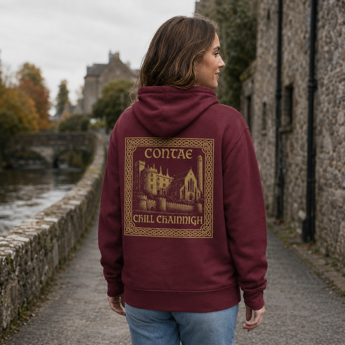 Woman wearing the maroon Kilkenny Heritage Hoodie from the back on a quiet medieval riverside lane